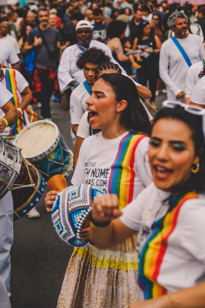 Integrantes de um coletivo de percussão com camisetas de arco-íris cantando no cortejo
